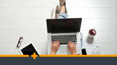 Widok z góry na zorganizowane stanowisko pracy osoby pracującej na laptopie, otoczoną kawą, tabletem, kawa i słuchawkami na jasnej podłodze